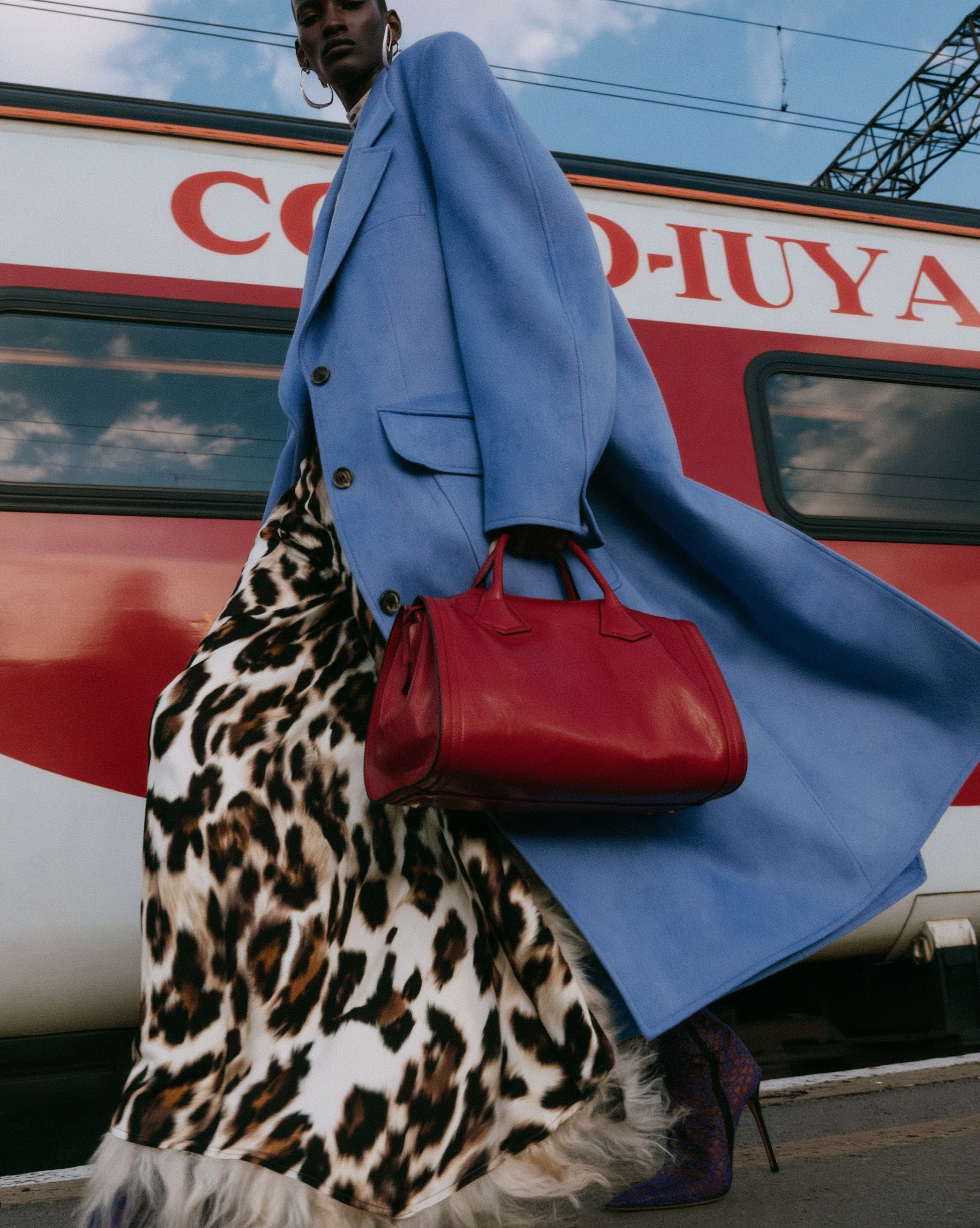 In Transit — Low-angle shot of a person in a blue coat and leopard-print skirt carrying a red handbag beside a train