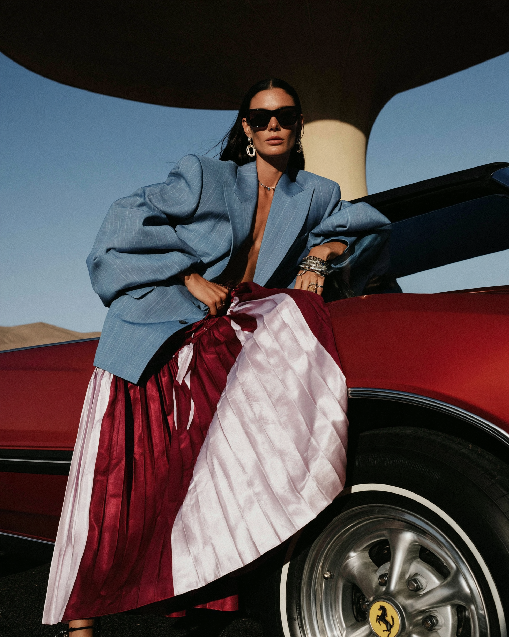 In Transit — Woman wearing sunglasses, oversized light blue blazer, and pleated satin skirt leaning against a classic red car outdoors