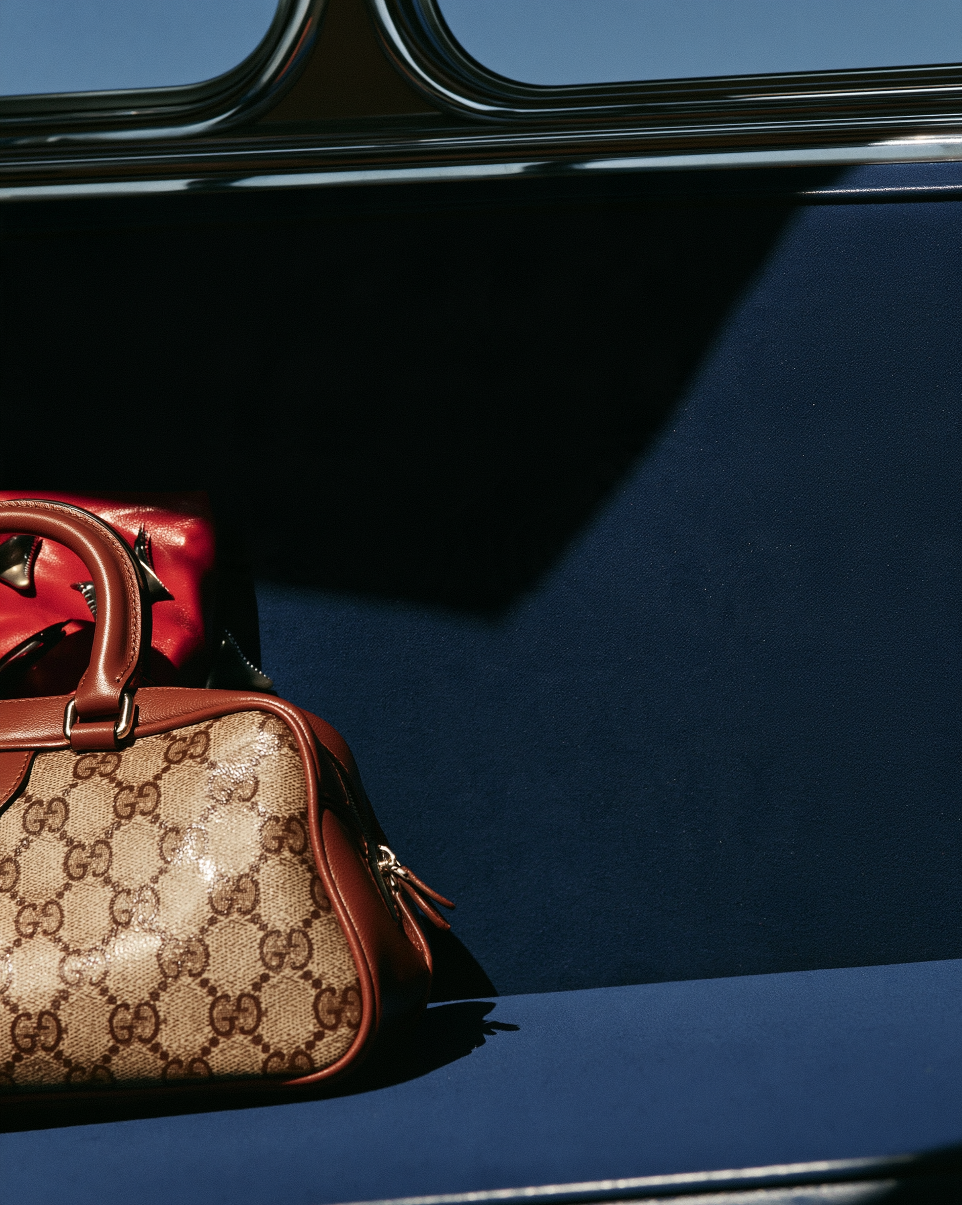 In Transit — A beige monogram handbag with brown leather trim and a red bag on a blue train seat in sunlight.