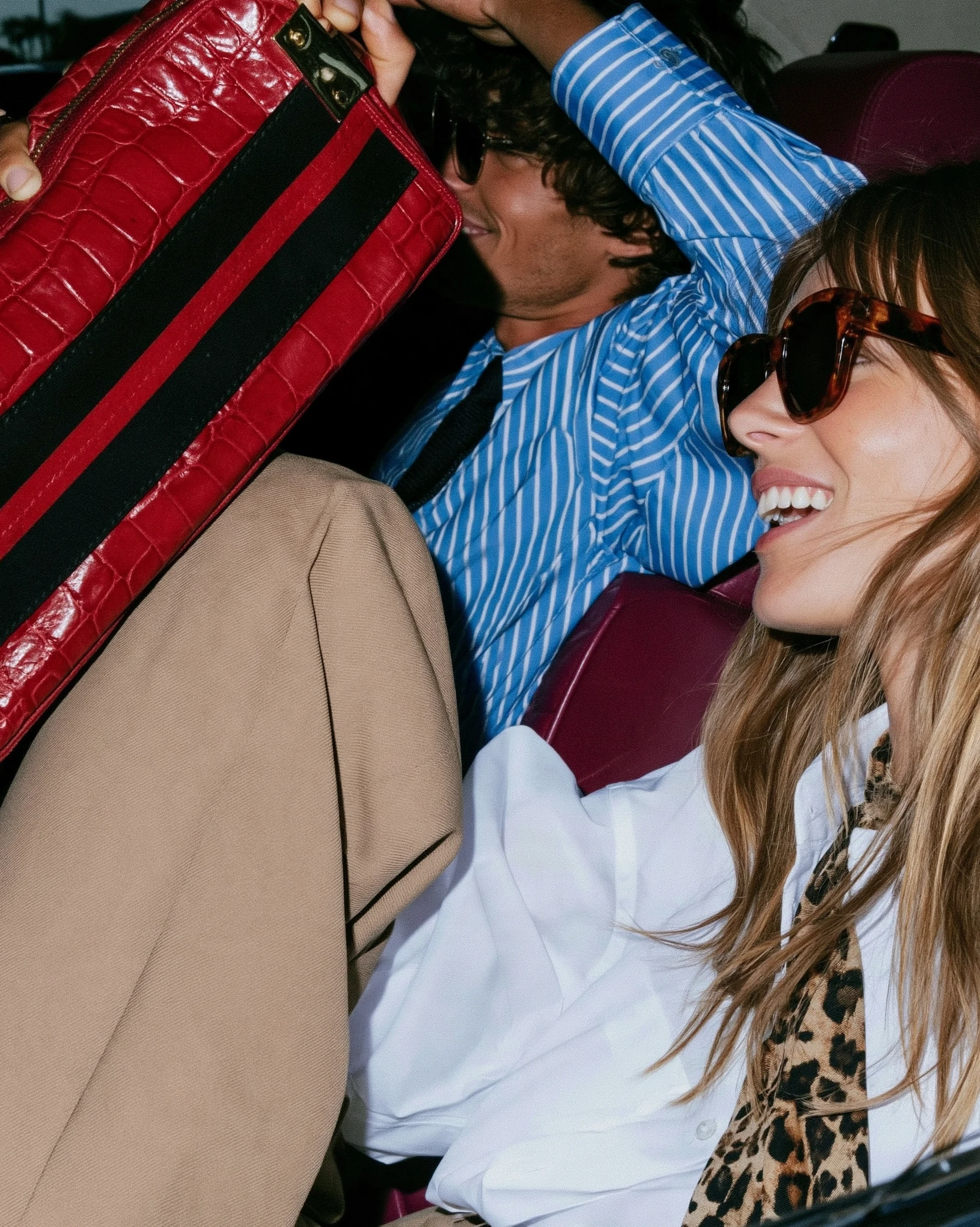 In Transit — Smiling woman in sunglasses and white shirt seated in a car beside a man holding a red bag