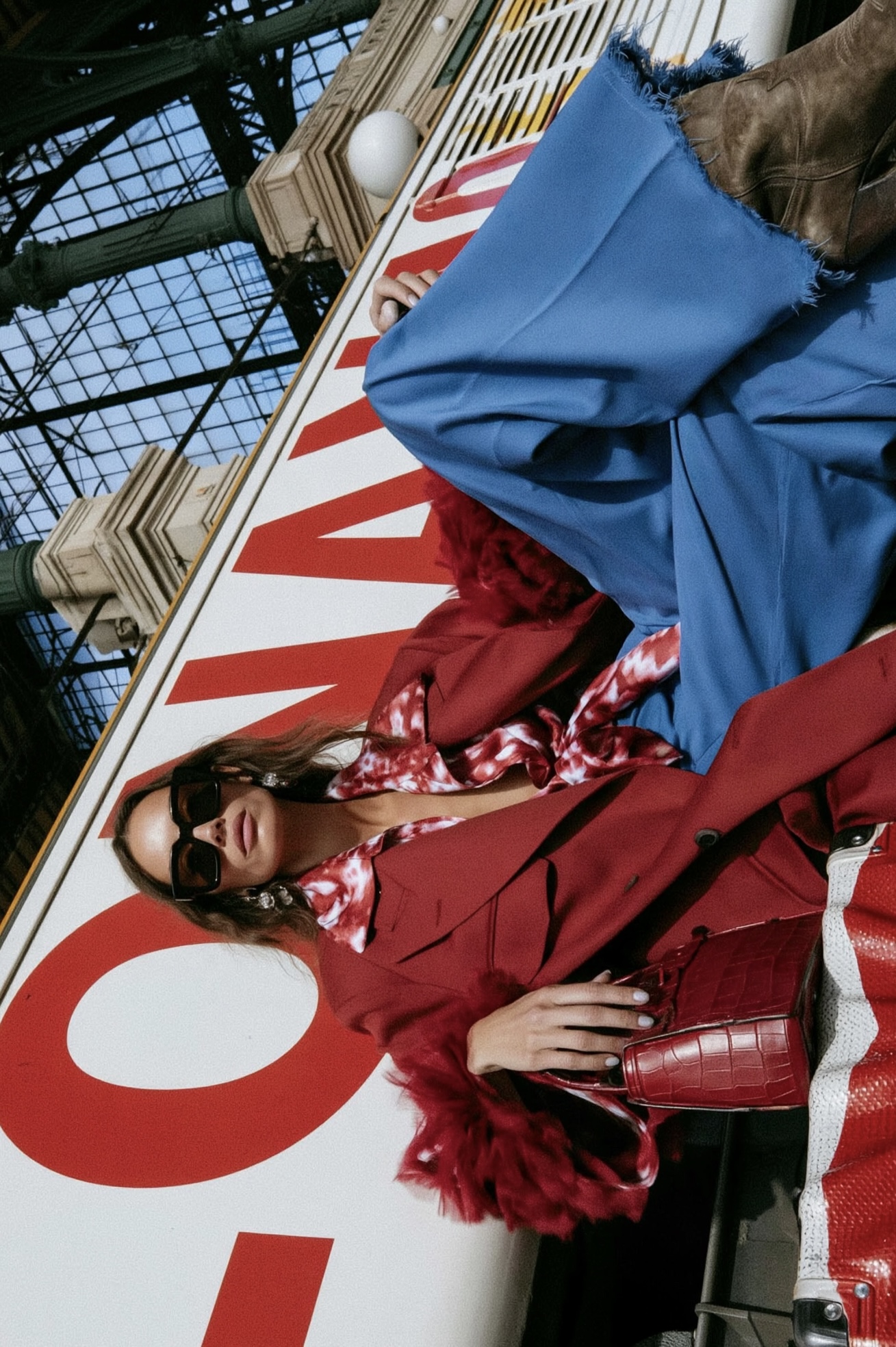 In Transit — Tilted portrait of a woman in sunglasses and a red blazer with blue trousers, brown boots and a red handbag beneath a station glass roof.