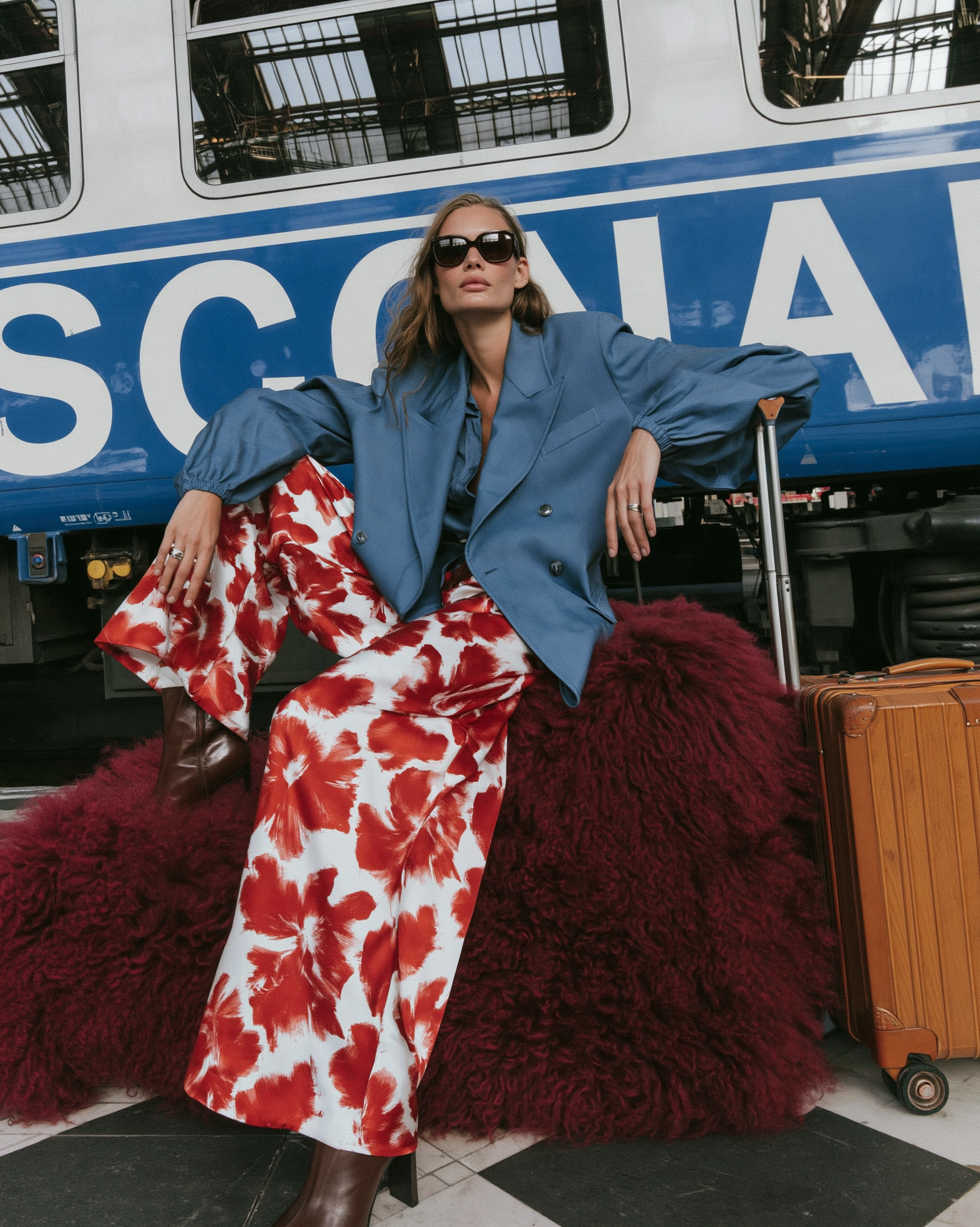 In Transit — A woman in sunglasses, a blue blazer and red-and-white floral trousers seated on burgundy shaggy fabric beside a train and tan suitcase.