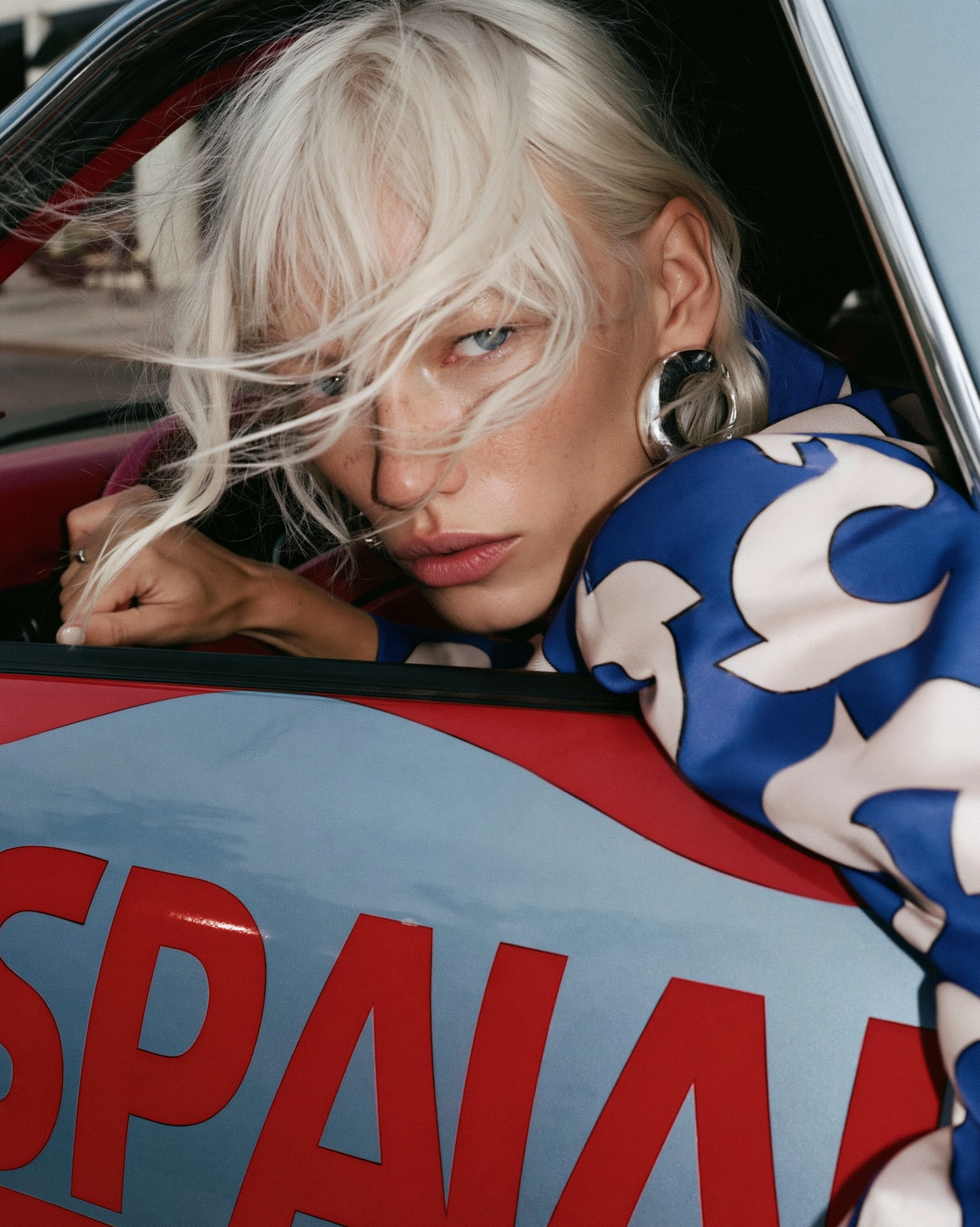 In Transit — Close-up of a blonde person in a blue patterned top leaning out a red and blue car window