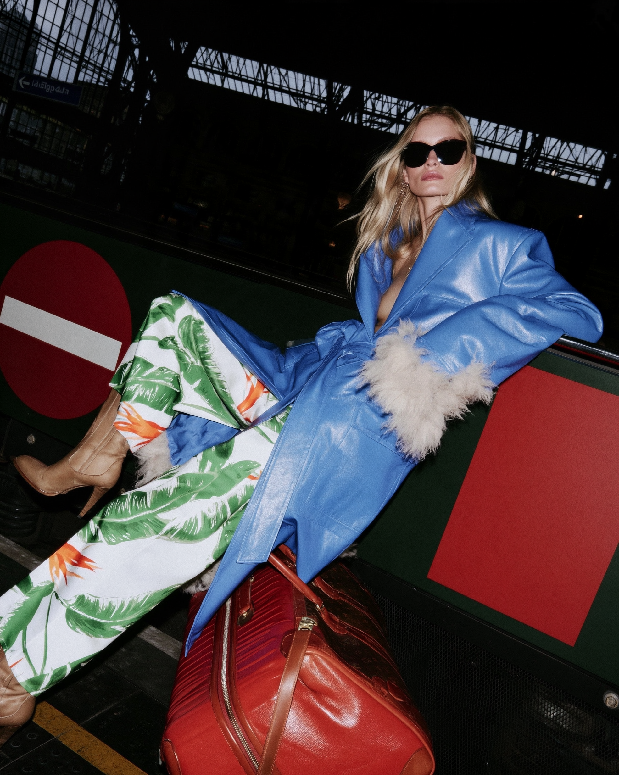 In Transit — Blonde woman in sunglasses, blue fur-trimmed coat, and palm-print pants reclining on red luggage at a train station