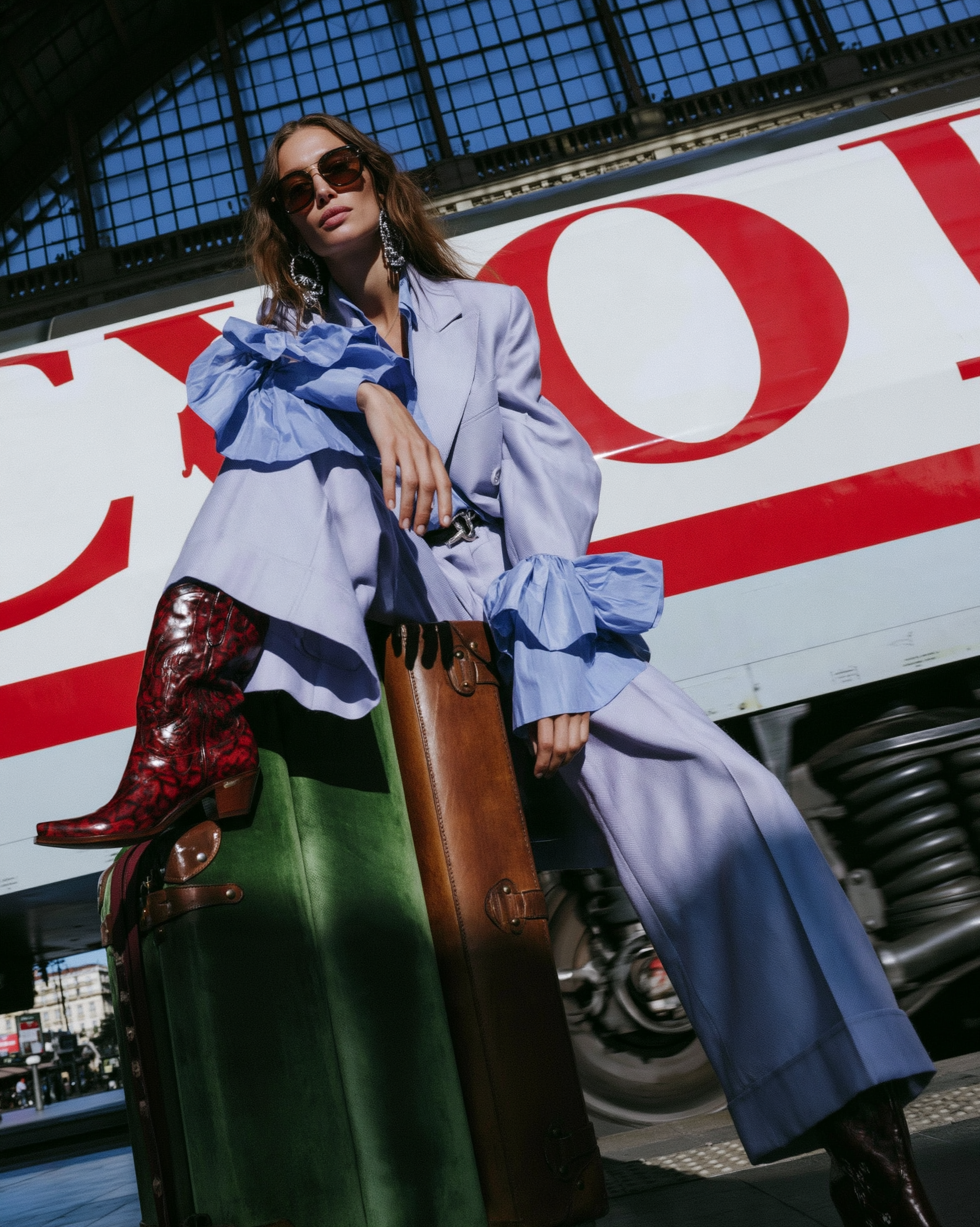 In Transit — Woman wearing a lavender ruffled suit and red boots seated on vintage suitcases at a train station