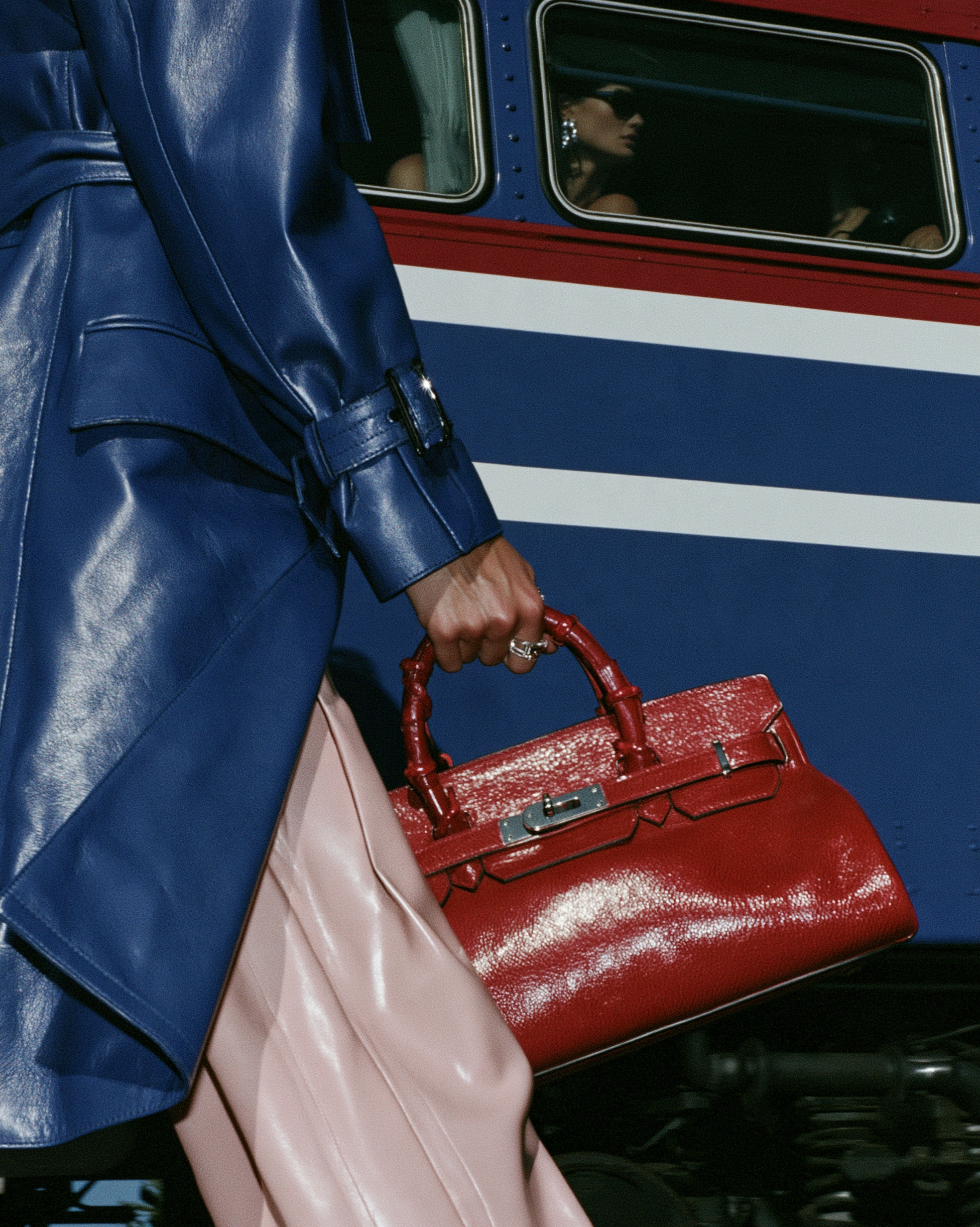 In Transit — Close-up of a blue leather coat and pale pink trousers with a glossy red handbag beside a blue, white and red train.